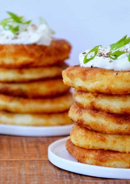 Stacks of mashed potato pancakes on white plates and a small bowl of scallions