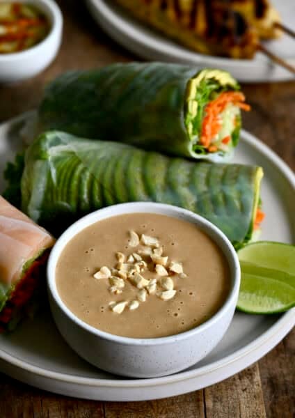 Thai peanut sauce in a bowl topped with chopped peanuts on a plate with fresh spring rolls.