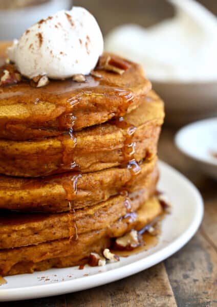 A tall stack of pumpkin pie pancakes topped with pecans, whipped cream and maple syrup.